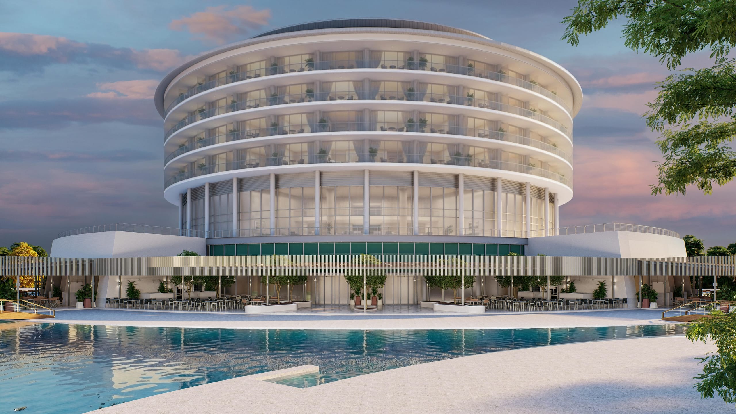 Dusk view of the round resort building with curved balconies, its main restaurant terrace under a slatted canopy at ground level, reflected in the pool in front