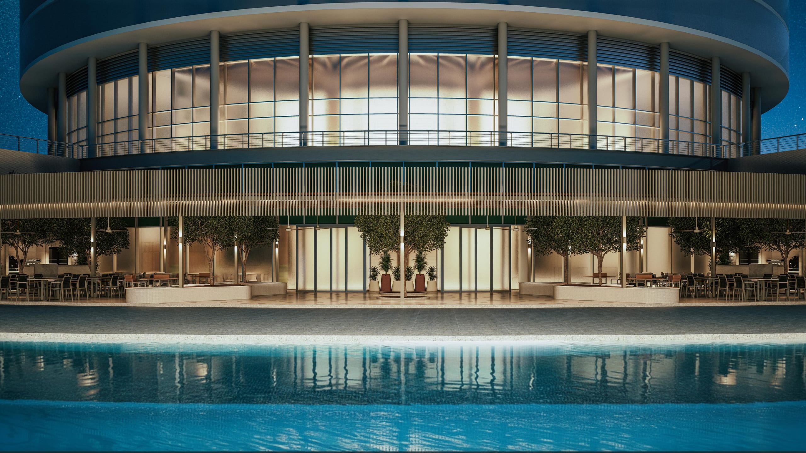 Night view across the pool to the glowing glazed ground floor of the round building and its terrace.