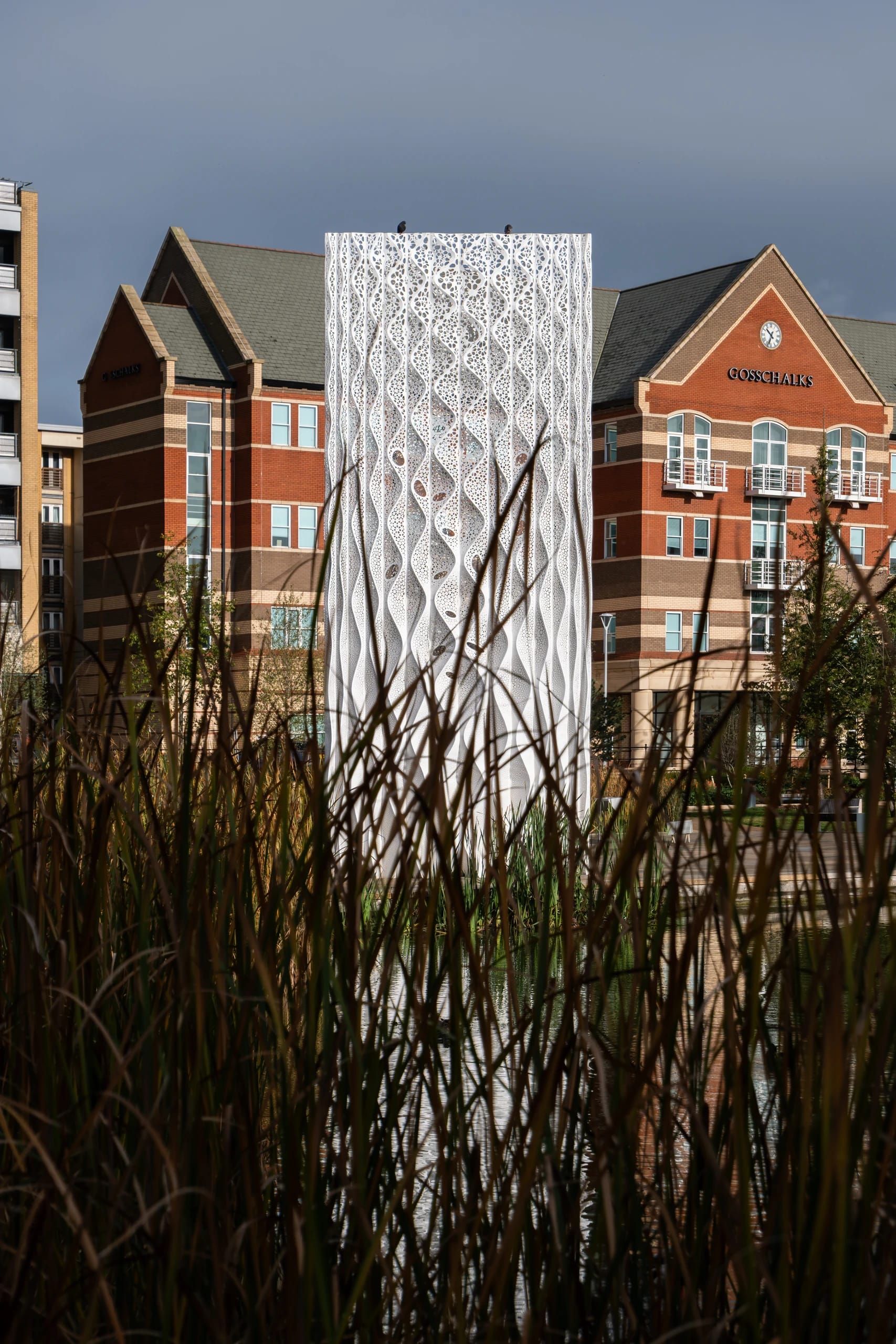 A white perforated cylindrical structure framed by reeds and brick buildings